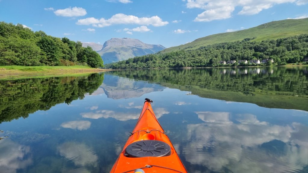View from orange kayak on calm lake with trees, mountains, houses and blue sky reflection.