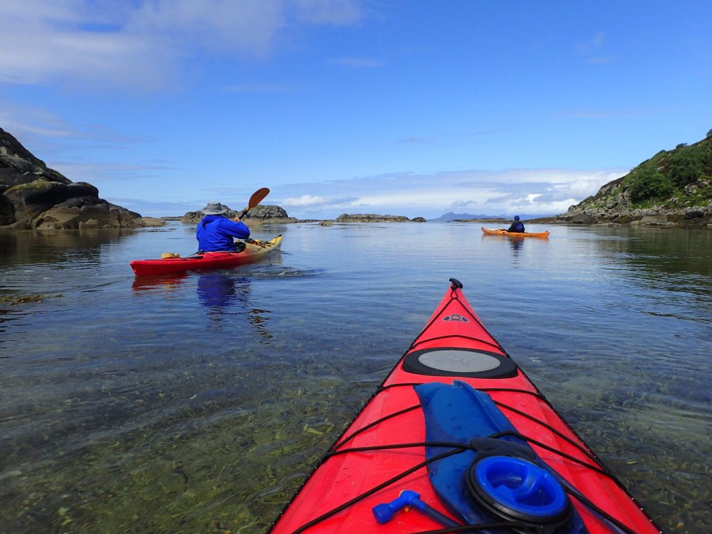 View from a red kayak of two people kayaking in calm coastal waters with rocky shores.